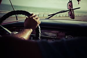 Picture of a man with his hands on steering wheel. Shades hanging in rear mirror.